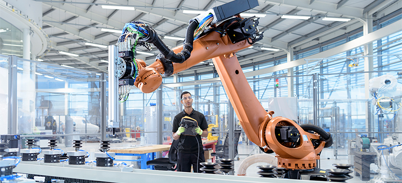 Egy ipari környezetben/gyárban dolgozó fiatalember egy nagy robotkart irányít.