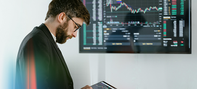 Egy szakember pénzügyi adatokat elemez egy táblagépen, miközben mögötte egy nagy kijelző piaci grafikonokat, árfolyamokat és kereskedési adatokat mutat — a számvitel és pénzügy szektor megjelenítése.