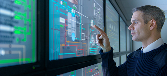 Egy szakember egy nagy digitális vezérlőpanellel lép interakcióba, amely összetett műszaki diagramokat és rendszeradatokat jelenít meg — a technológiai és mérnöki szektor ábrázolása.