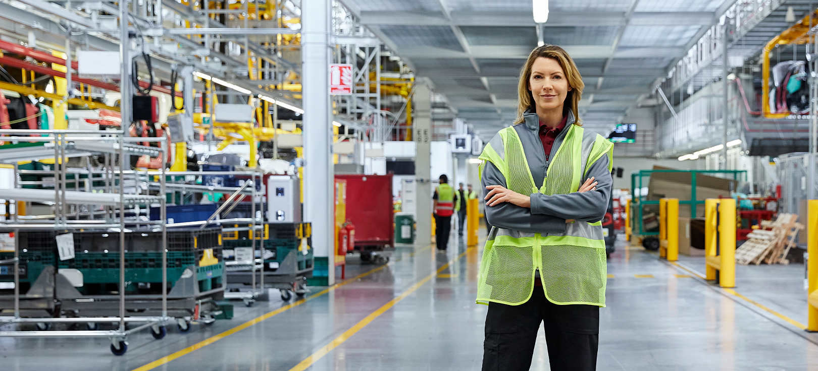 Magas láthatósági mellényt viselő személy áll összefont karral egy nagy, modern gyártóüzemben, ipari berendezések, gépek és gyártósorok között.