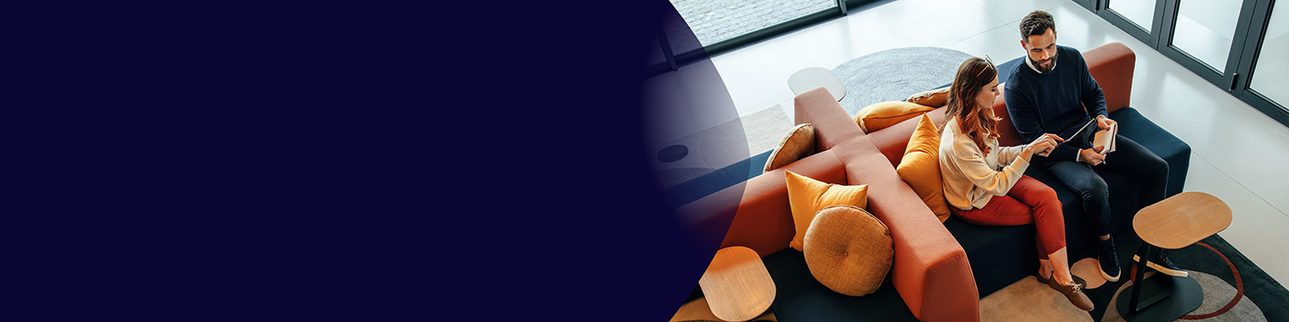 A man and woman sitting together in a modern office lounge area, looking at a tablet while seated on colourful sofas with round cushions and small side tables nearby.