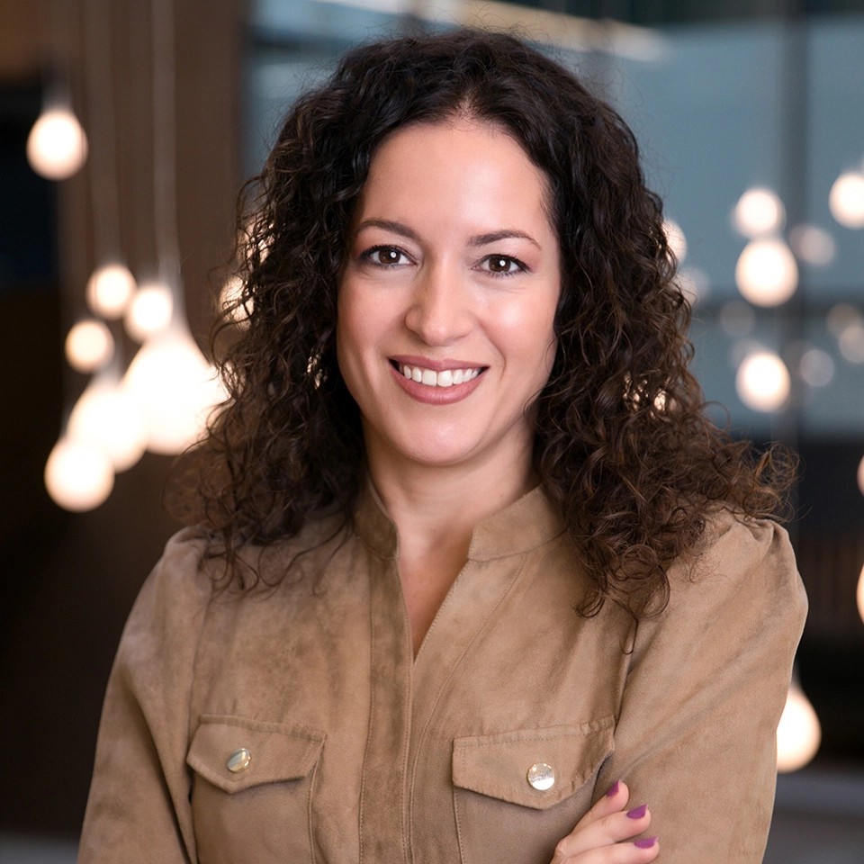 Göndör, sötét hajú nő semleges színű blúzban, beltérben, elmosódott, meleg fényű háttér előtt.